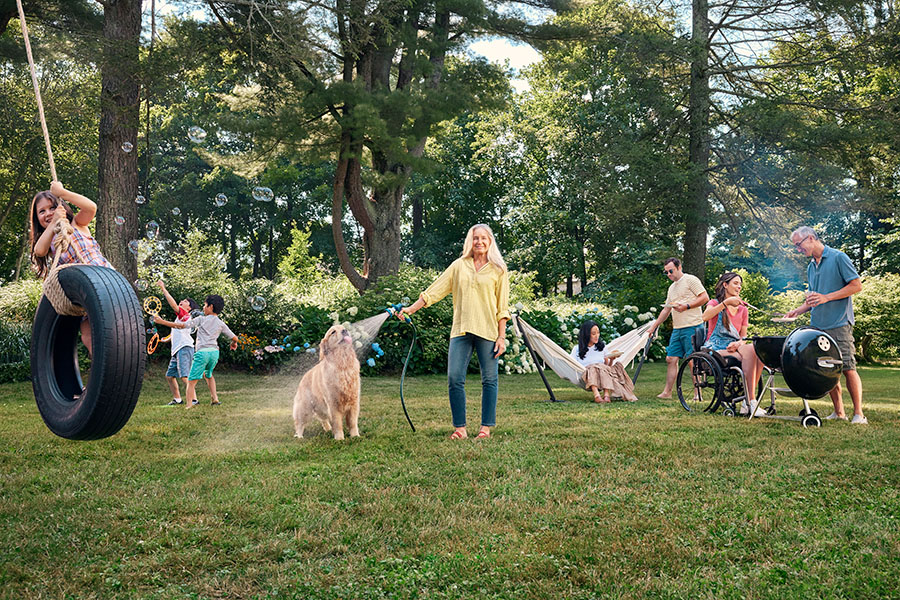A full backyard, a woman waters the lawn while a dog drinks from the hose, children play on a tire swing and chase each other, and others gather around a grill.